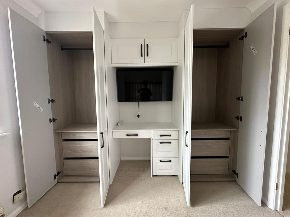 Custom white shaker bedroom storage unit with open oak-lined wardrobes, built-in desk, drawers, and wall-mounted television.