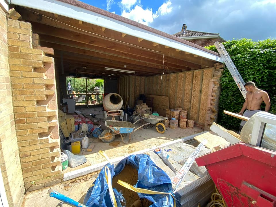 Garage conversion in progress with timber stud walls, exposed roof joists, cement mixer, and stacked bricks.