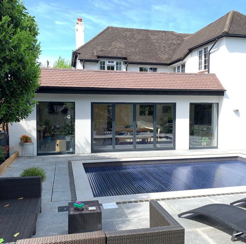 Completed white-rendered rear house extension with bifold doors, stone patio, swimming pool, and outdoor seating on a sunny day.