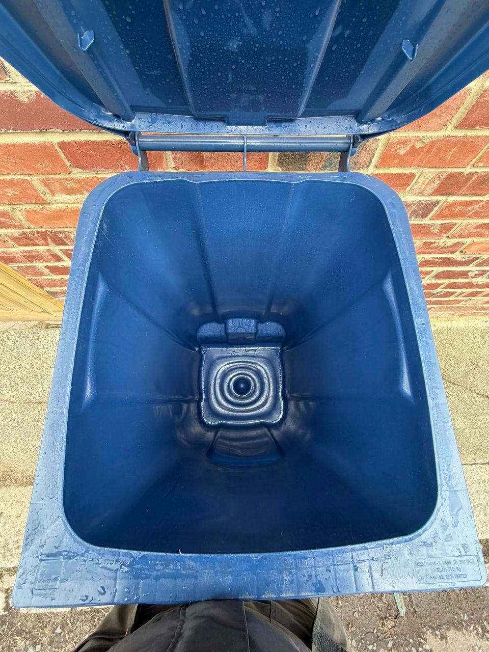Freshly cleaned blue recycling wheelie bin with open lid and water droplets, against a brick wall