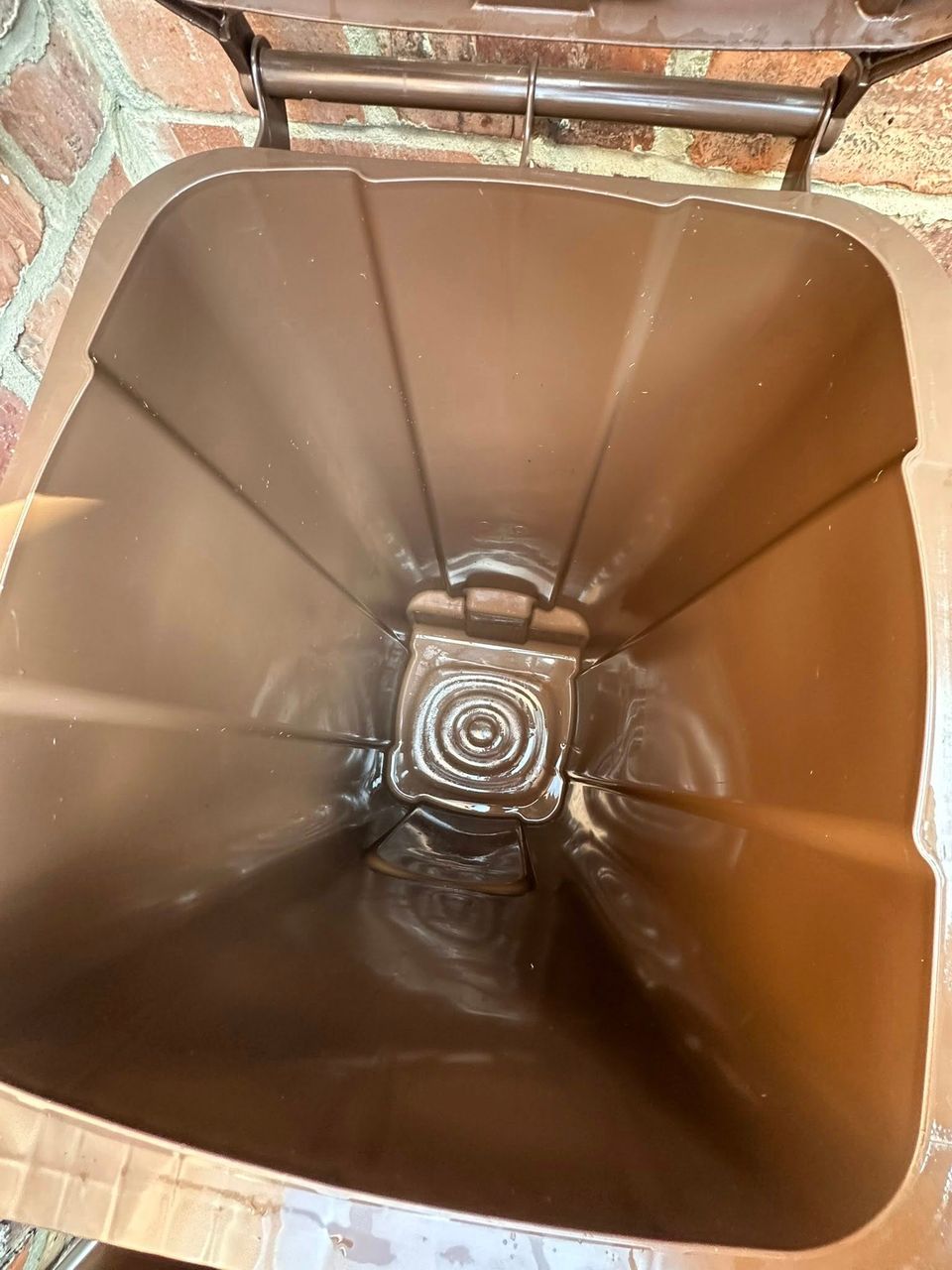 Freshly cleaned brown garden-waste wheelie bin with glossy interior against a red brick wall